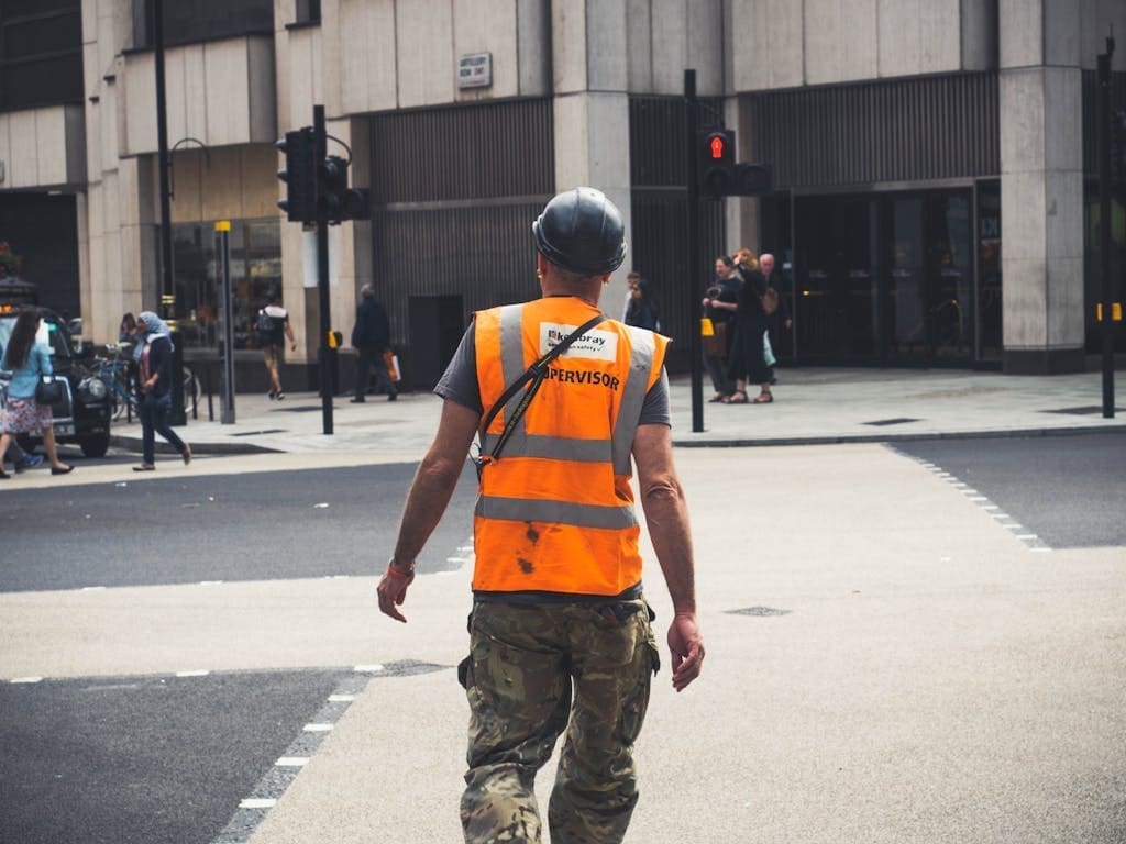 Back view of unrecognizable young male constructor in protective helmet and vest walking on asphalt road in modern city district